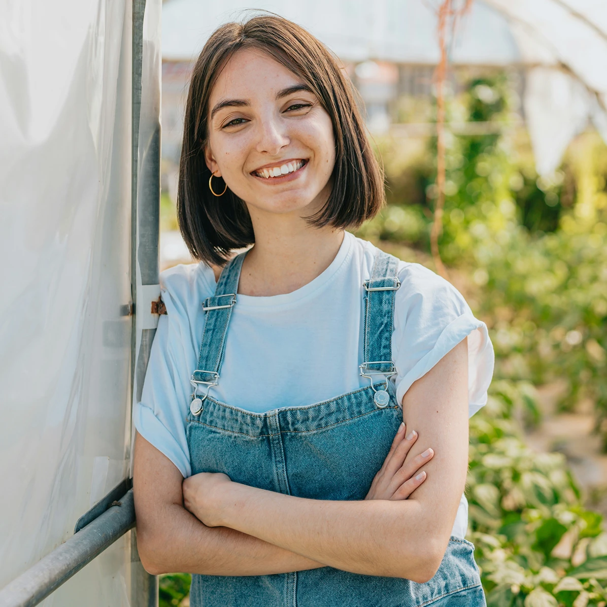 A young woman standing in a greenhouse with her arms crossed smiling
