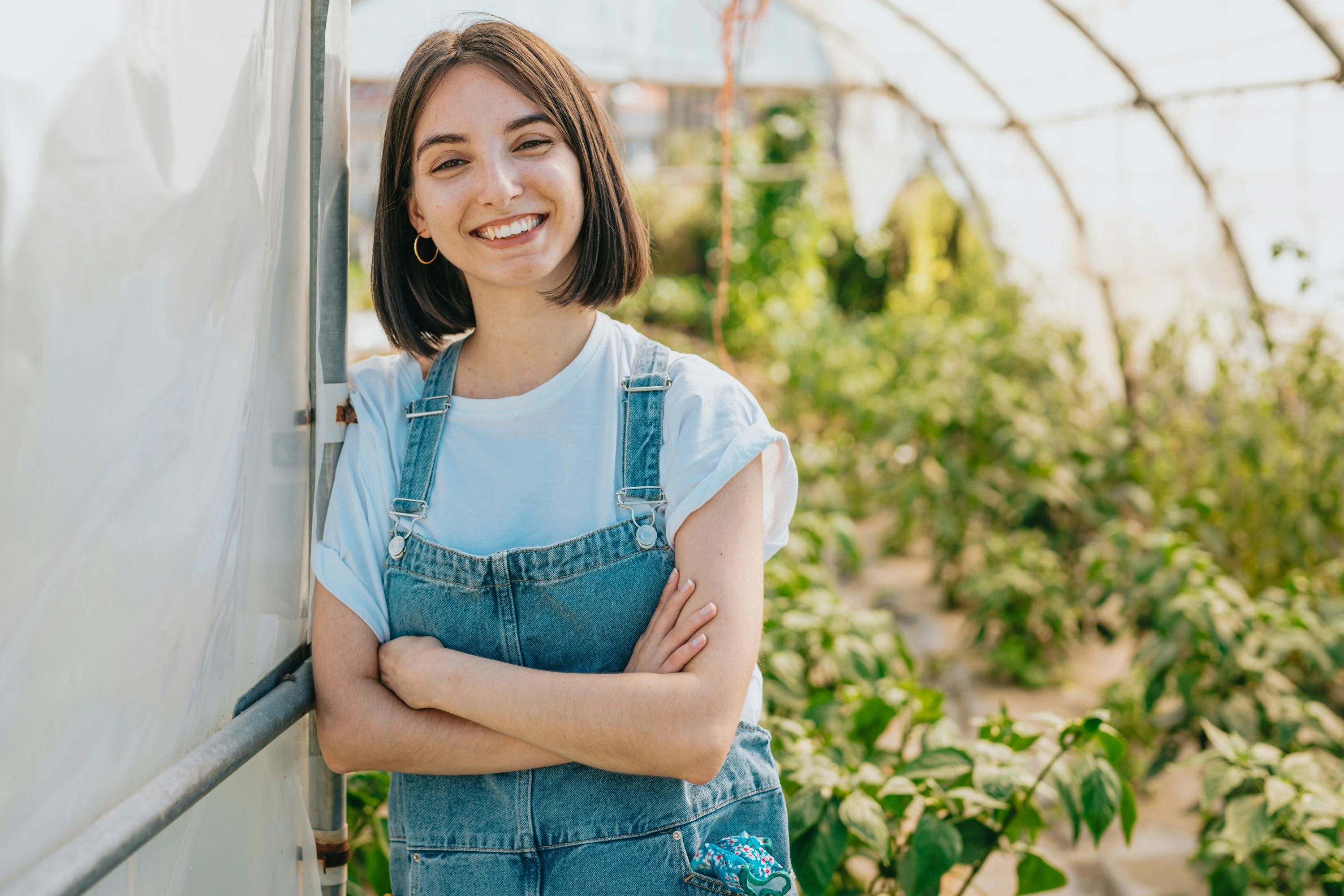 A young woman standing in a greenhouse with her arms crossed smiling