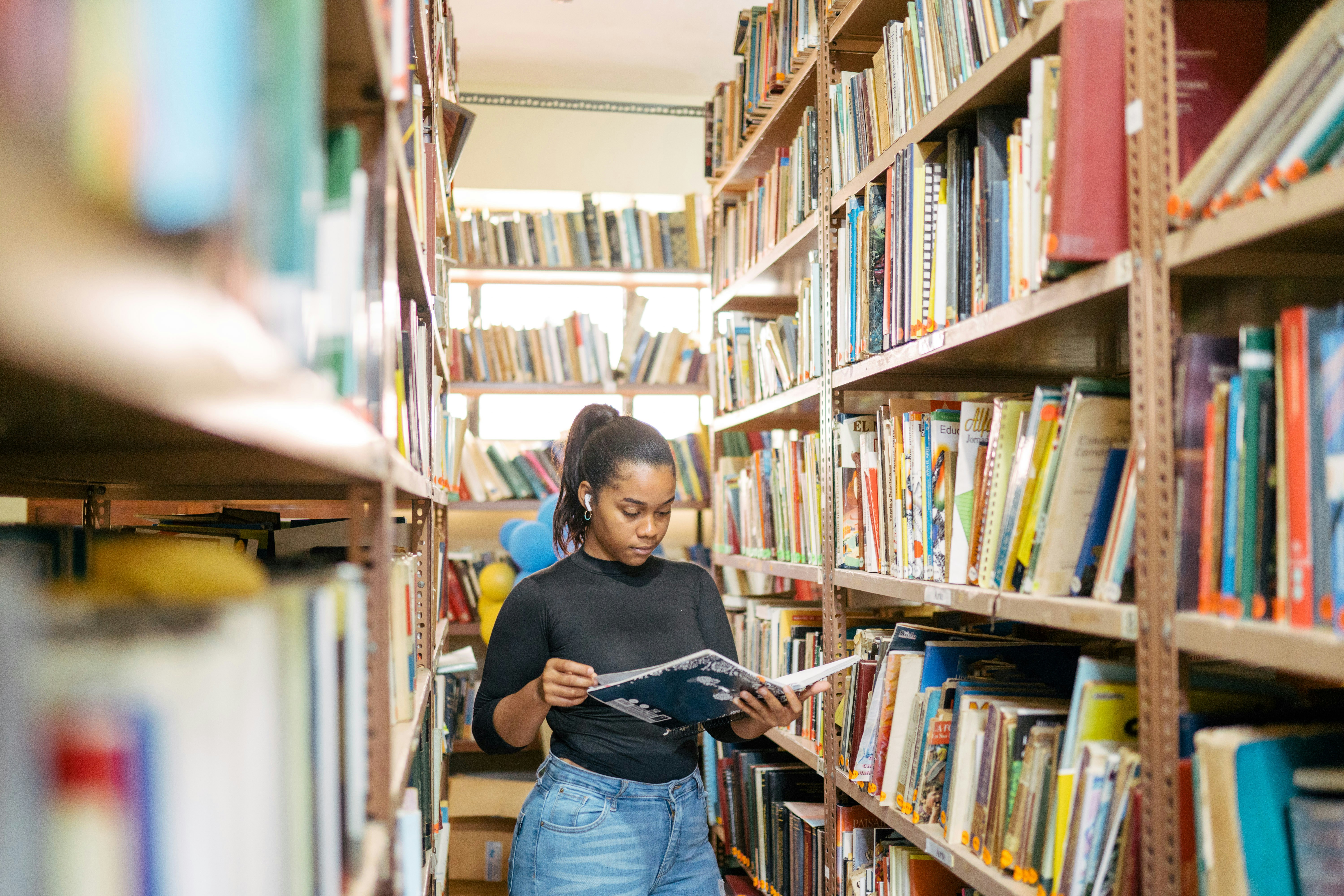 A young woman is reading a book in a library, shelves of books surround her