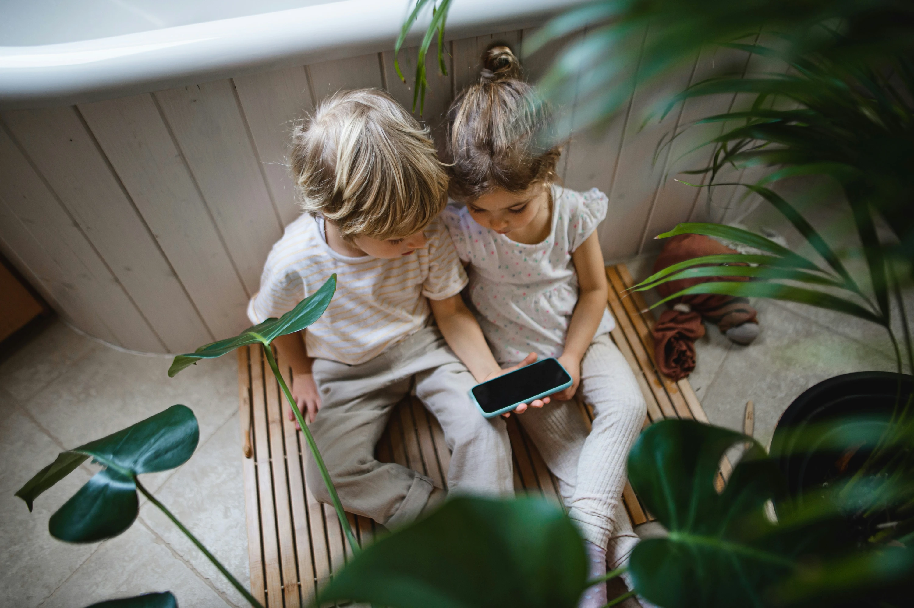 Two small children are sitting on floor indoors, using a smartphone