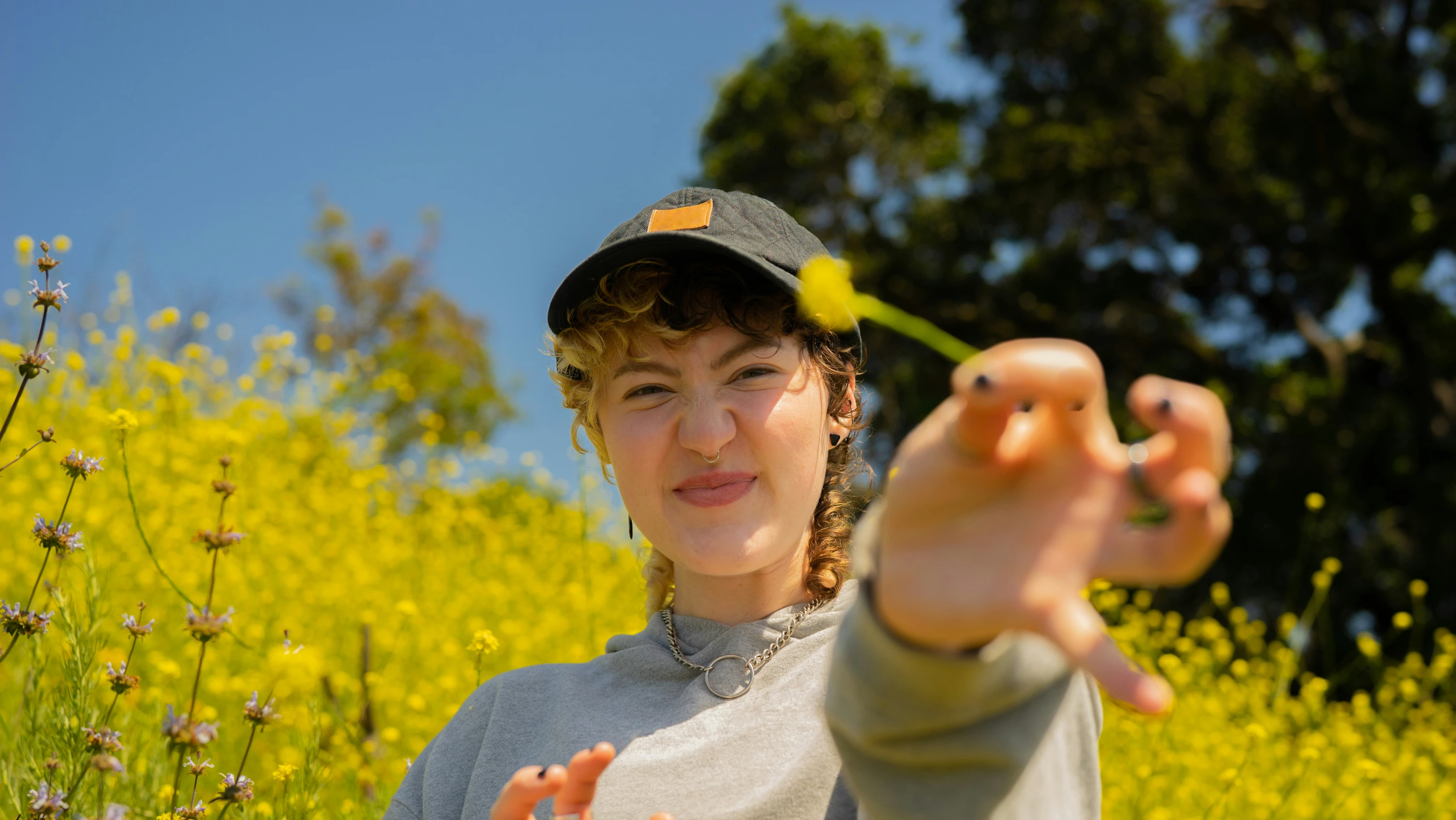 A young woman is smiling for the camera whilst stood outside on a sunny day holding a daisy in her hand
