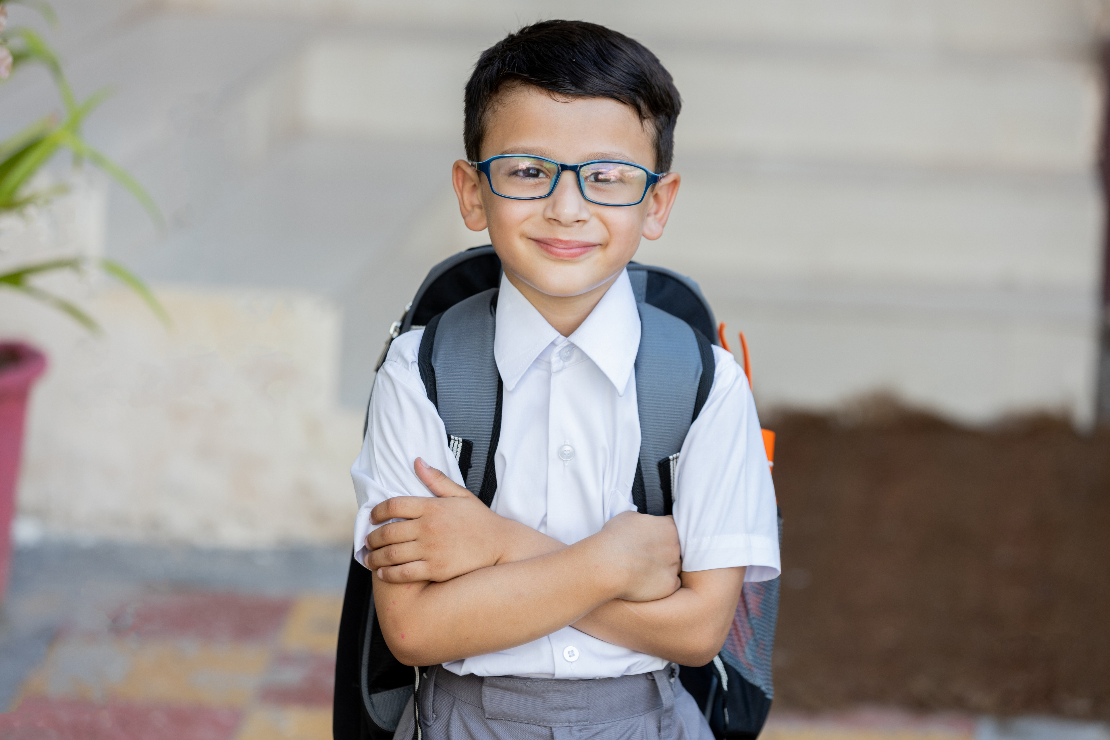 A young boy with glasses is smiling at the camera whilst wearing a school uniform and backpack