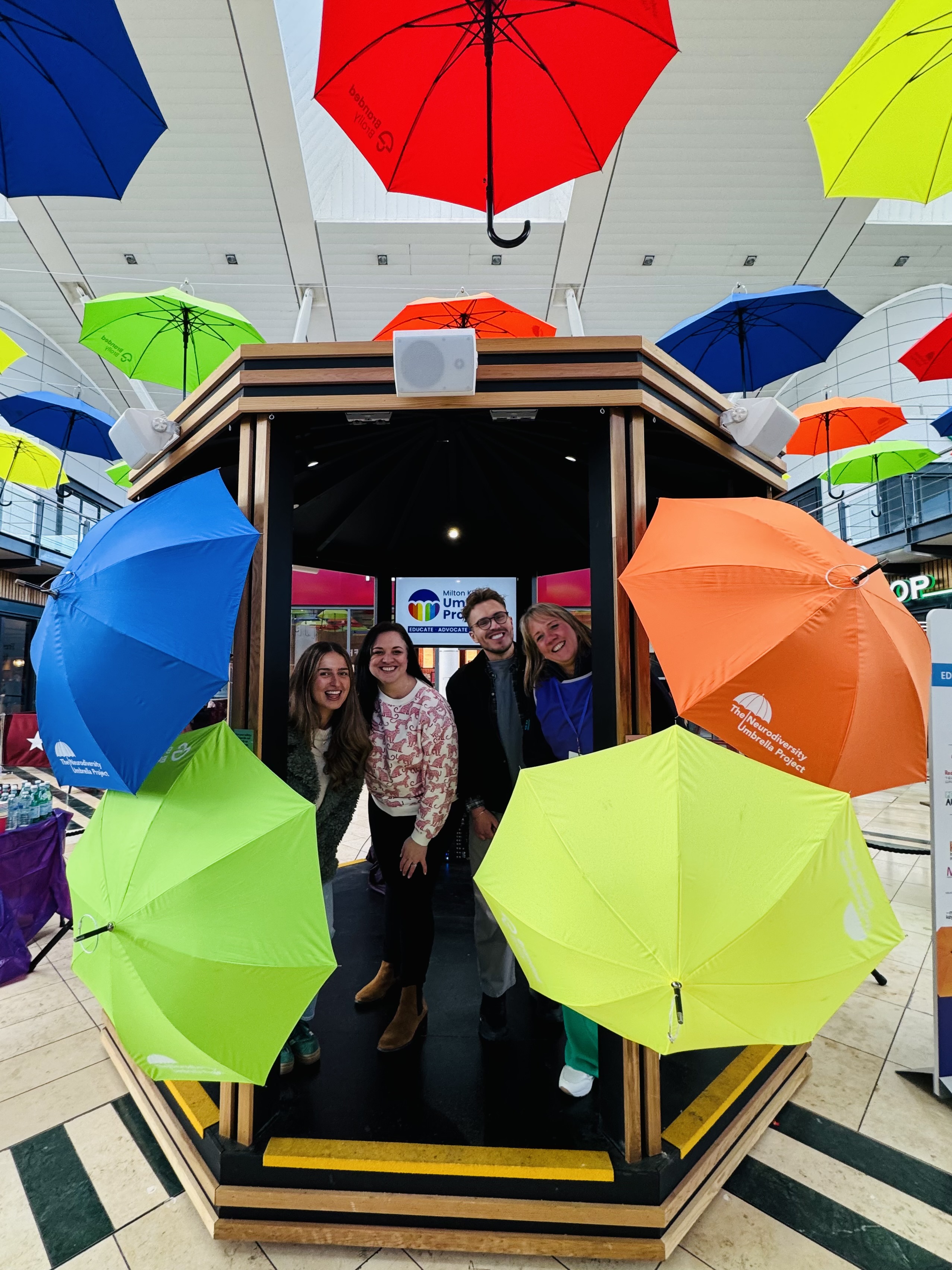 3 members of the ADHD Foundation and Jane Lambert from Makewell MK posing with umbrellas by the Midsummer Place Umbrella Project display