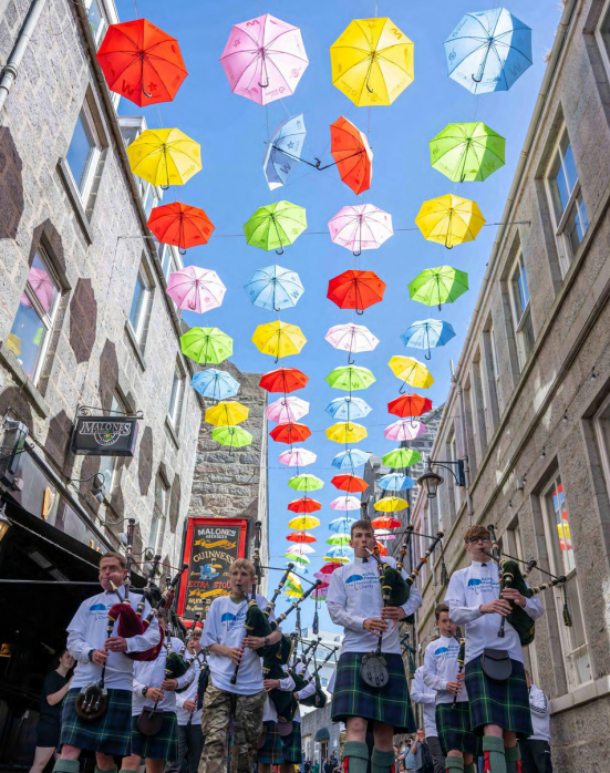 Group of young men playing the bagpipes under the outdoor umbrella project display at Shiprow Village, Aberdeen